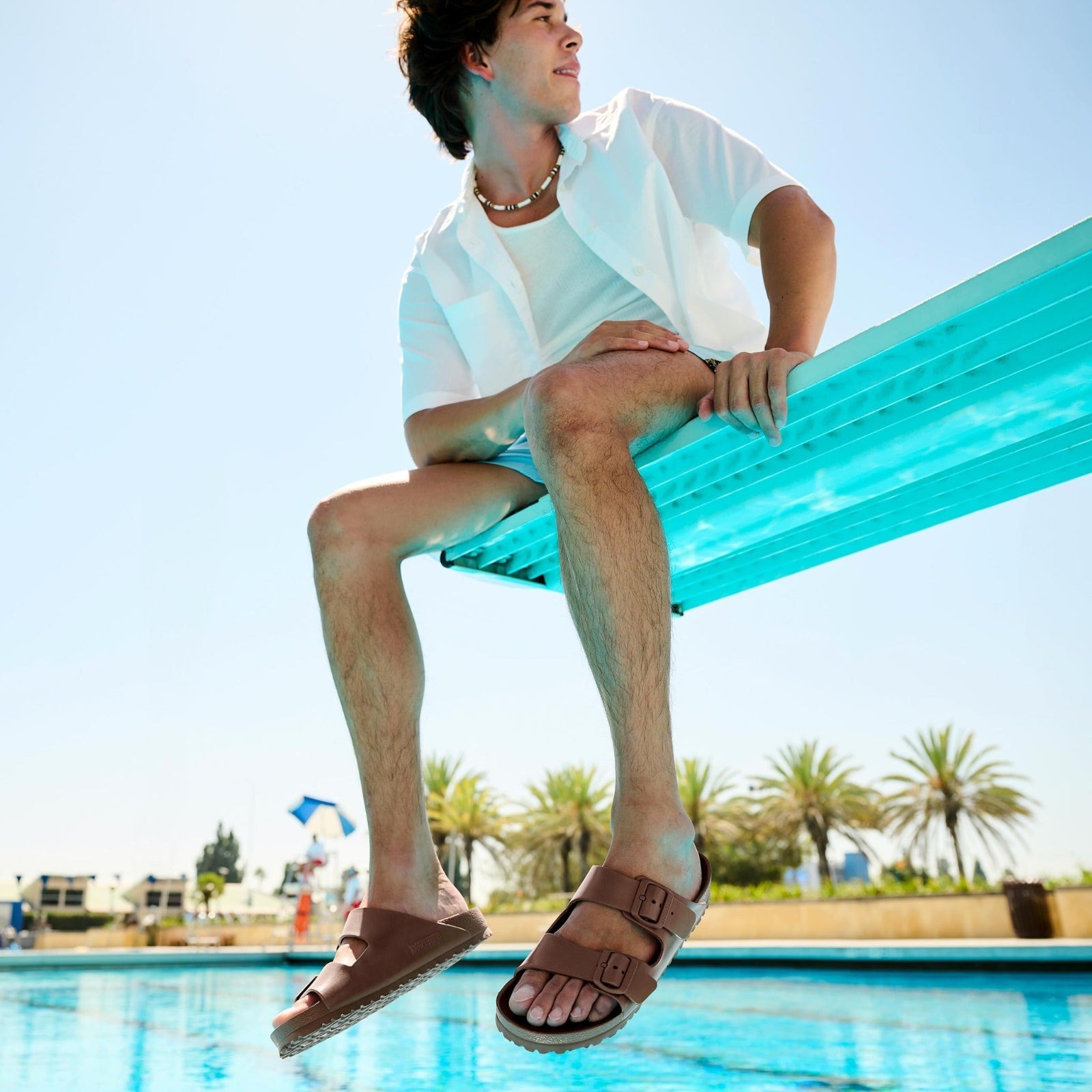 Men sitting on a blue inflatable pool float by a pool with palm trees in the background.