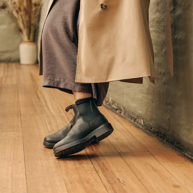 View of Chisel Toe 068 Black Boot Blundstone Canada worn by female model wearing black boots and a beige coat on a wooden floor.