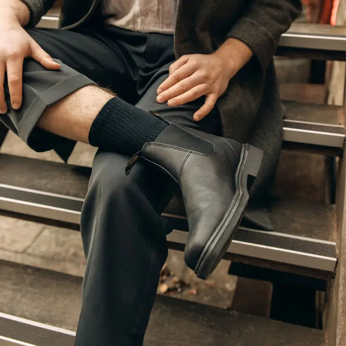 Person wearing black leather boots Blundstone Canada Chisel 068 and black socks sitting on a metal bench.