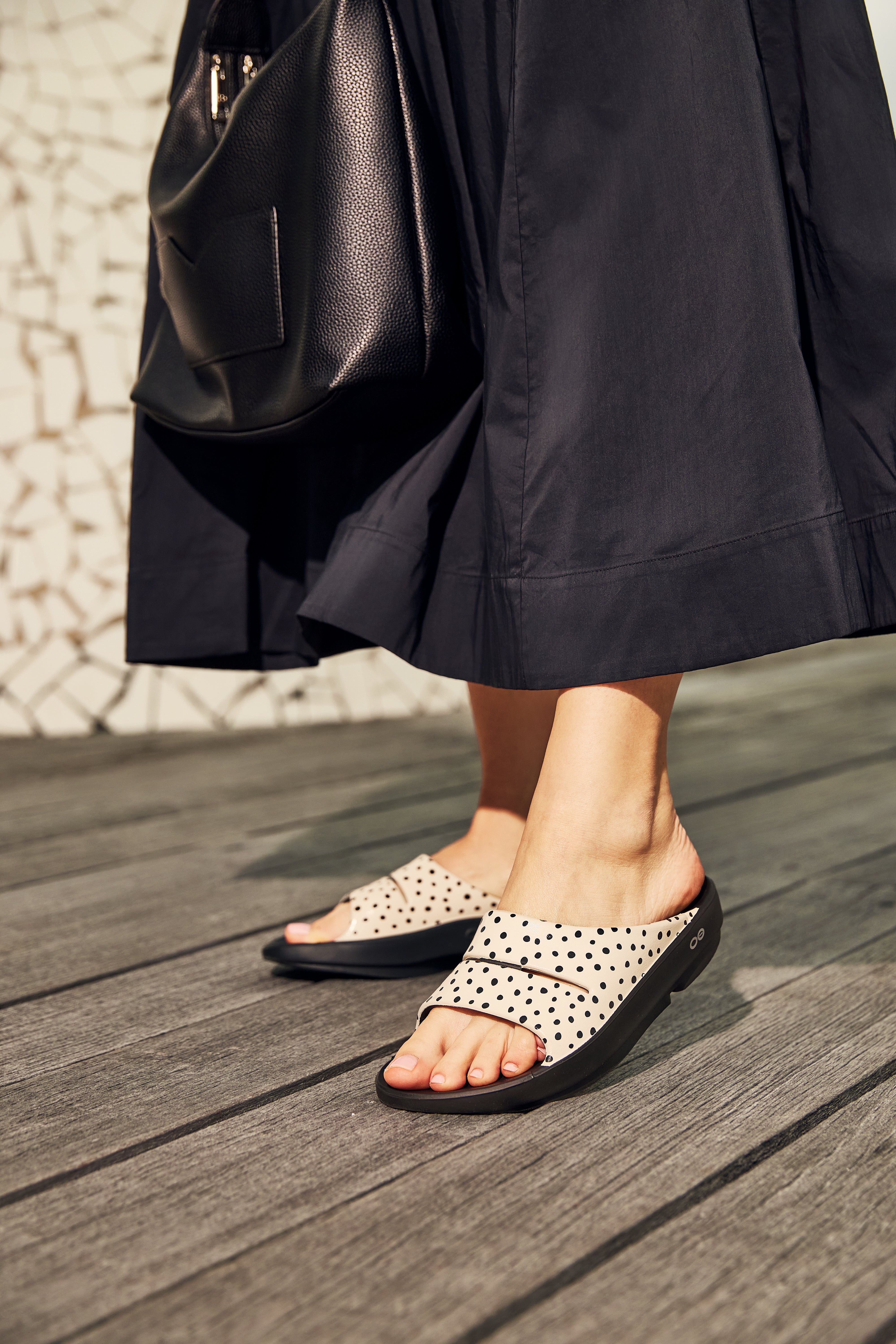 OOFOS OOahh Limited Pebble Slide Sandal woemne wearing dot sandals on a wooden floor with a black backpack.