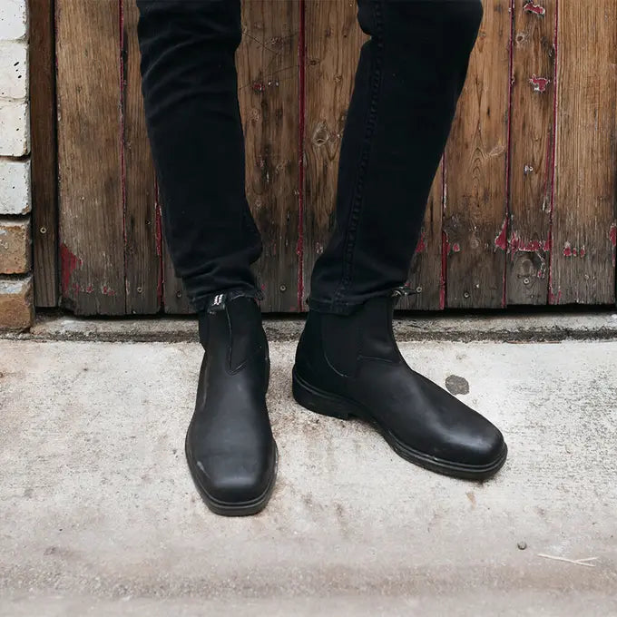 View of Chisel Toe 068 Black Boot Blundstone Canada worn by male model  with black pants against a wooden door background. 