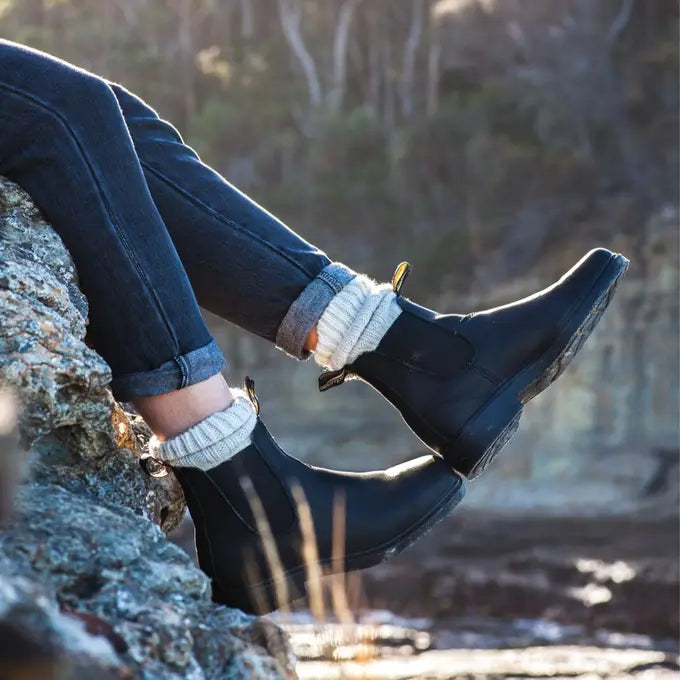 Person wearing black rubber boots Blundstone Canada Chisel 068 and white socks sitting on a rock with a natural background