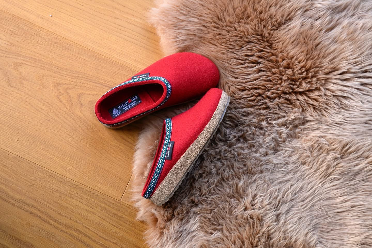 HAFLINGER  CLASSIC GRIZZLY SLIPPERS Red slippers with blue accents on a wooden floor next to a fluffy rug.