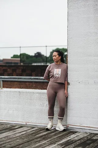 Female wearing a brown outfit leaning against a wall on a wooden deck NEW BALANCE WMORLB6 Fresh Foam X More v6 Shoes 