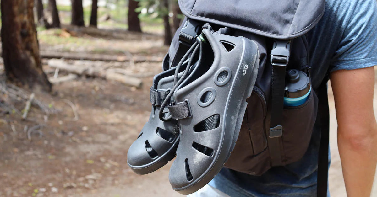 Person wearing a backpack with OOFOS OOCANDOO BLACK sandals attached, in a forest setting