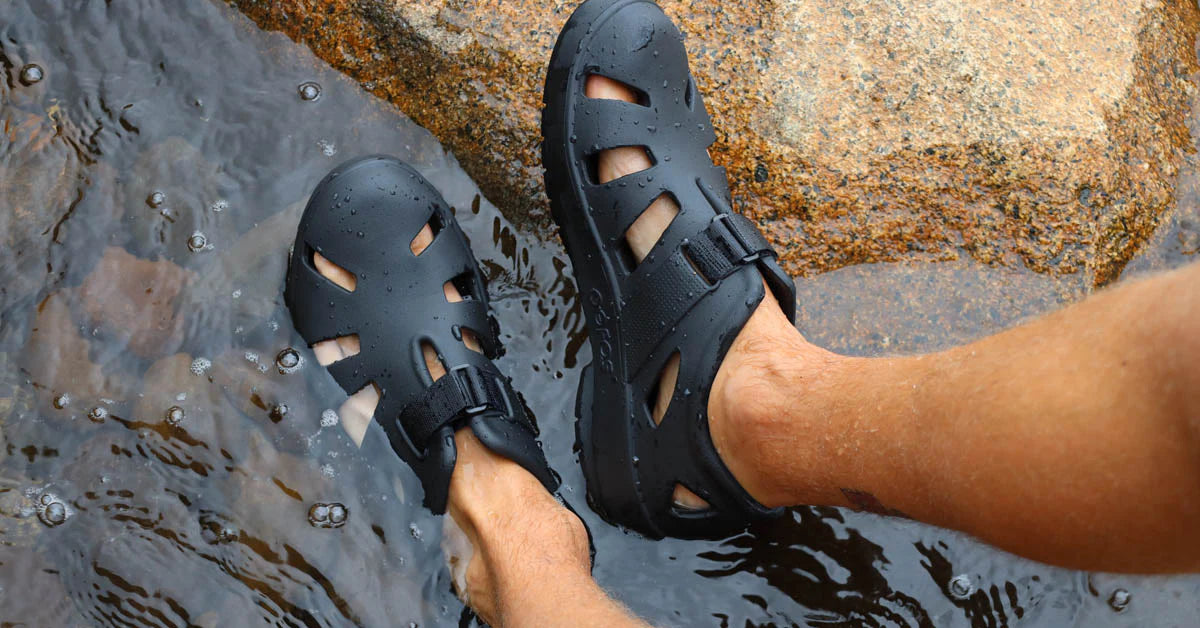 OOFOS OOCANDOO Black sandals worn on feet stepping into water near rocks